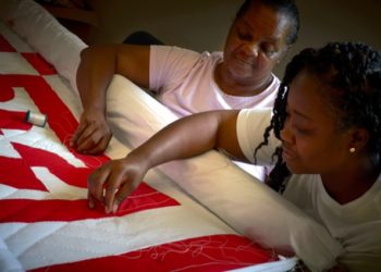 two black women bent over a quilt with a red pattern