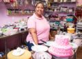 Woman in a pink shirt standing behind a counter of colorful cakes.